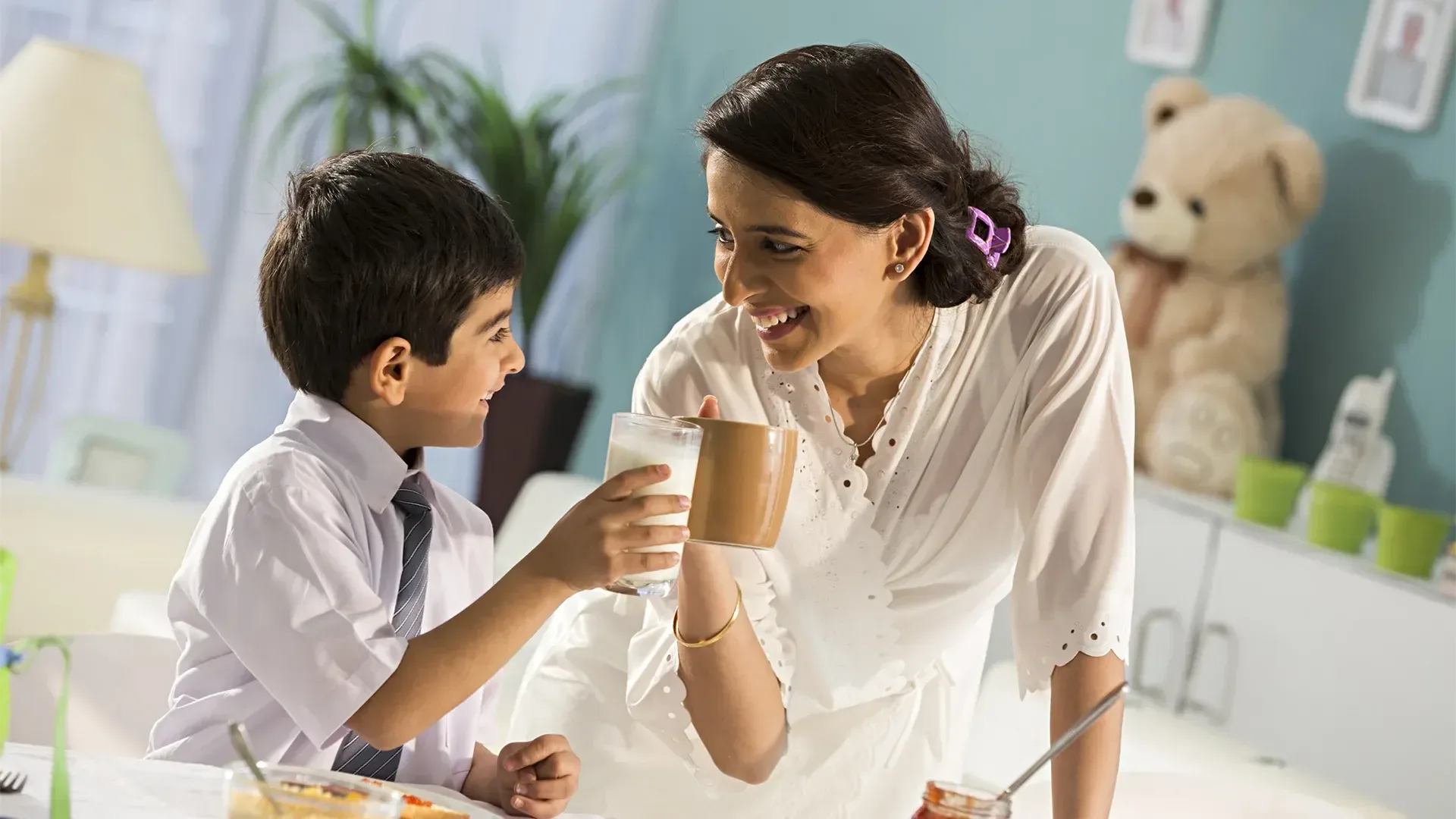 a smiling mother and her young son clinking glasses of milk and coffee at the breakfast table in a home setting